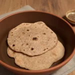 Stack of freshly cooked roti flatbreads on a wooden board with a skillet in background.